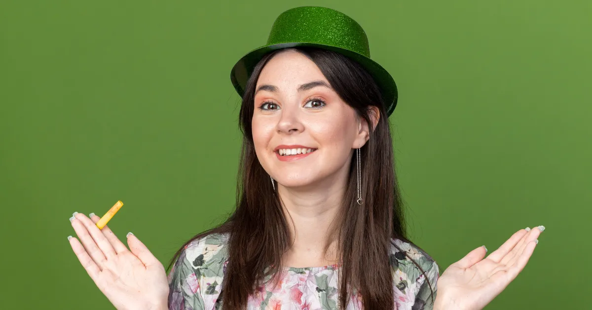 Smiling woman wearing a green glitter hat and floral top, holding a small yellow cosmetic item, posing against a green background.