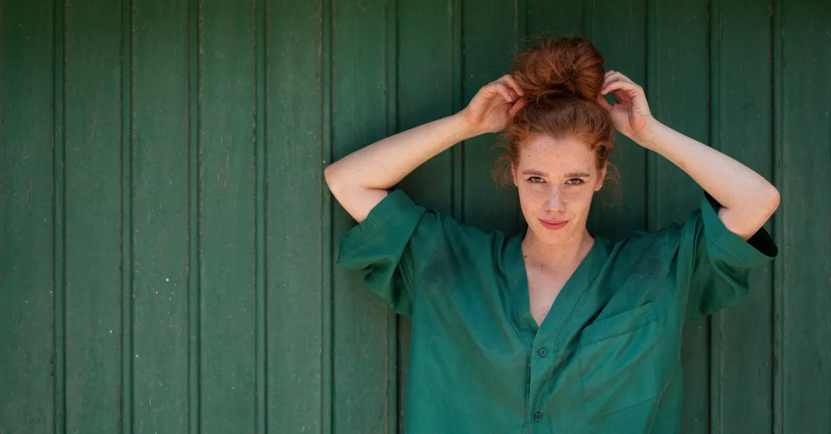 A woman wearing a green shirt stands in front of a green wooden wall, adjusting her hair in a bun and looking at the camera.