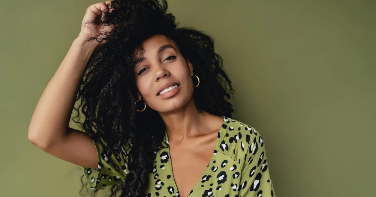 A woman with curly hair wearing a green patterned dress smiles softly while touching her hair against a green background.