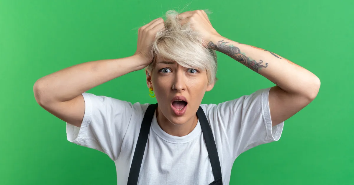 A woman with short blonde hair wearing a white shirt and black apron looks shocked while holding her head against a green background.