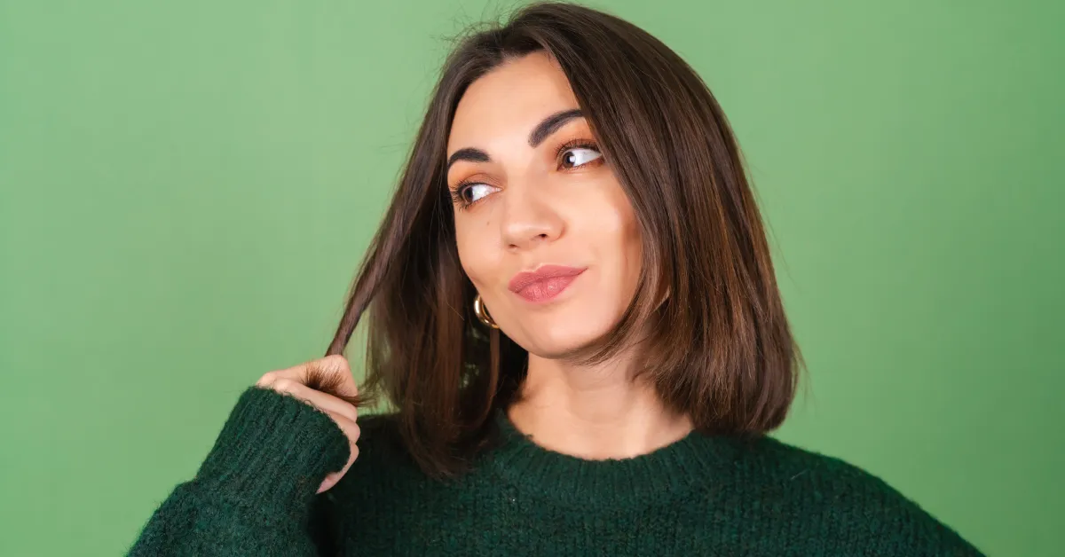 Woman with shoulder-length brunette hair wearing a dark green sweater against a light green background, showcasing a cozy fall hair color idea.
