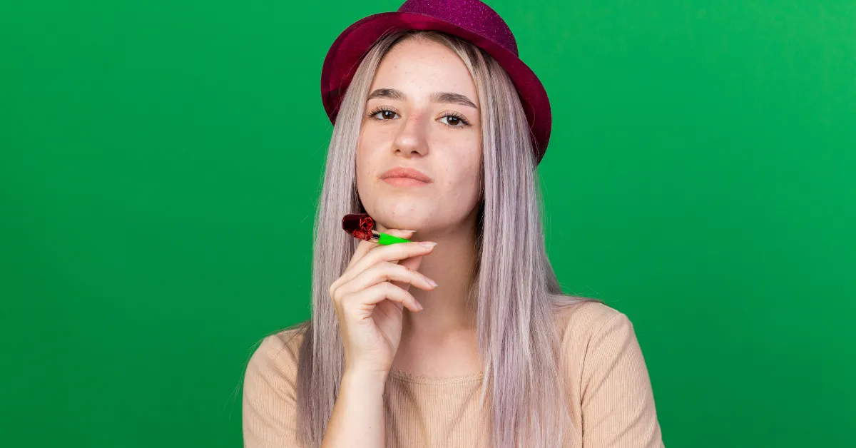 A young woman wearing a maroon hat stands against a green background, holding a small party horn near her chin and looking at the camera.