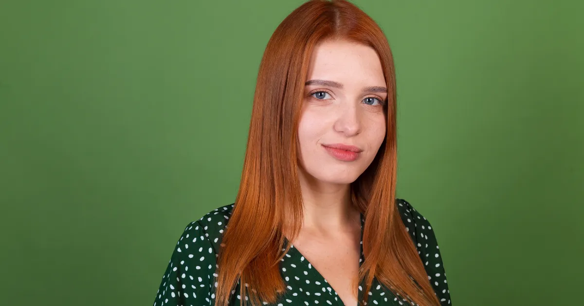 A young woman with long red hair wearing a green polka dot dress looks at the camera against a green background.