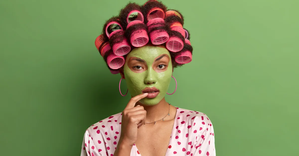 Woman wearing pink hair rollers and a green facial mask, dressed in a polka dot top, posing against a light green background.