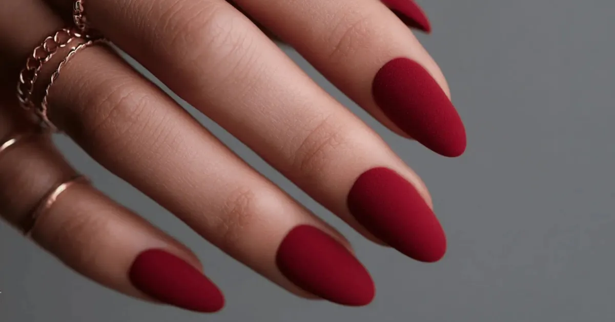 Matte red almond-shaped nails on a hand with delicate gold rings against a neutral background.