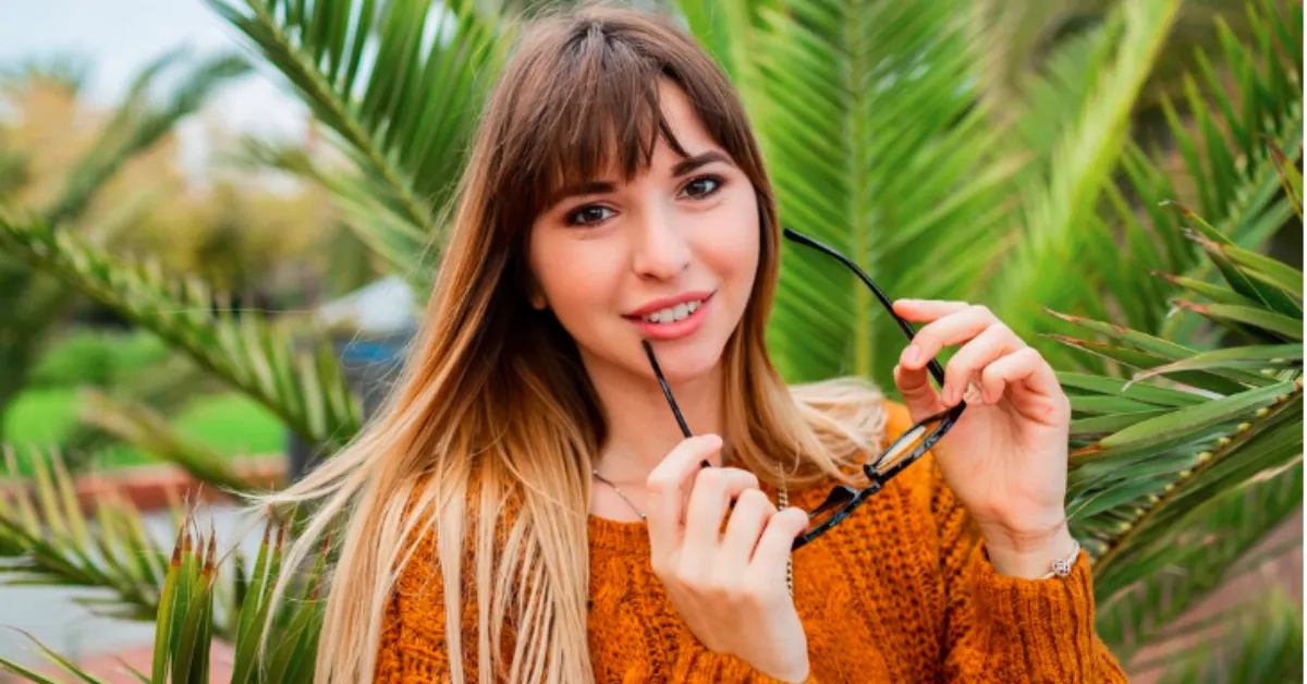 A smiling young woman wearing an orange sweater stands in front of palm leaves while holding a pair of sunglasses and looking at the camera