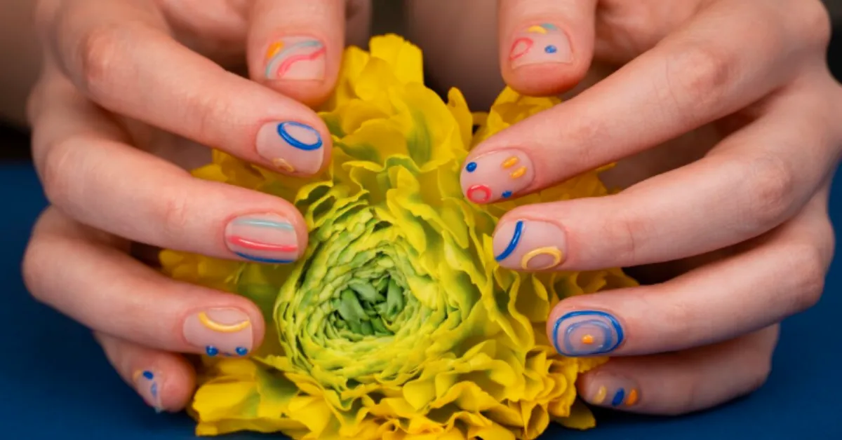Colorful abstract nail art wali ungliyon se ek bright yellow flower (ranunculus jaisa layered phool) ko gently pakda hua hai, neeche blue background dikh raha hai.