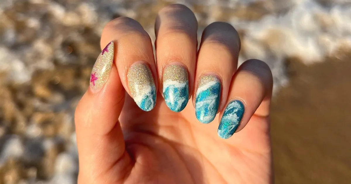Close-up of a hand with almond-shaped nails featuring ocean-inspired nail art,blue waves, white foam details, and glitter accents,held against a blurred beach background.